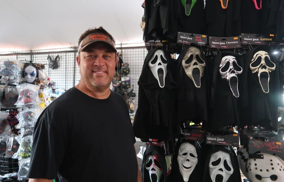 Robbie Wright, owner of Halloween Express Asheville, smiling in front of the Ghost Face mask wall inside the tent